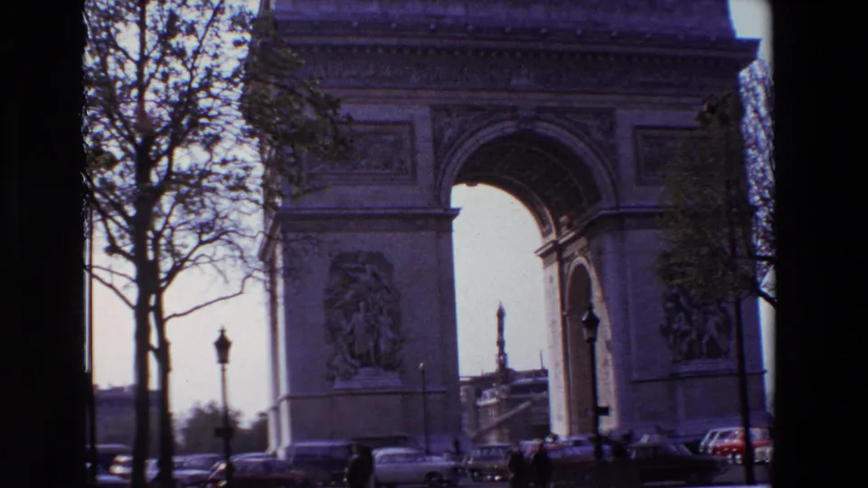 1969 Paris frame of the Arc de Triomphe with tree branches, street traffic, pedestrians, and the monument's sculptural facade visible in faded color film.