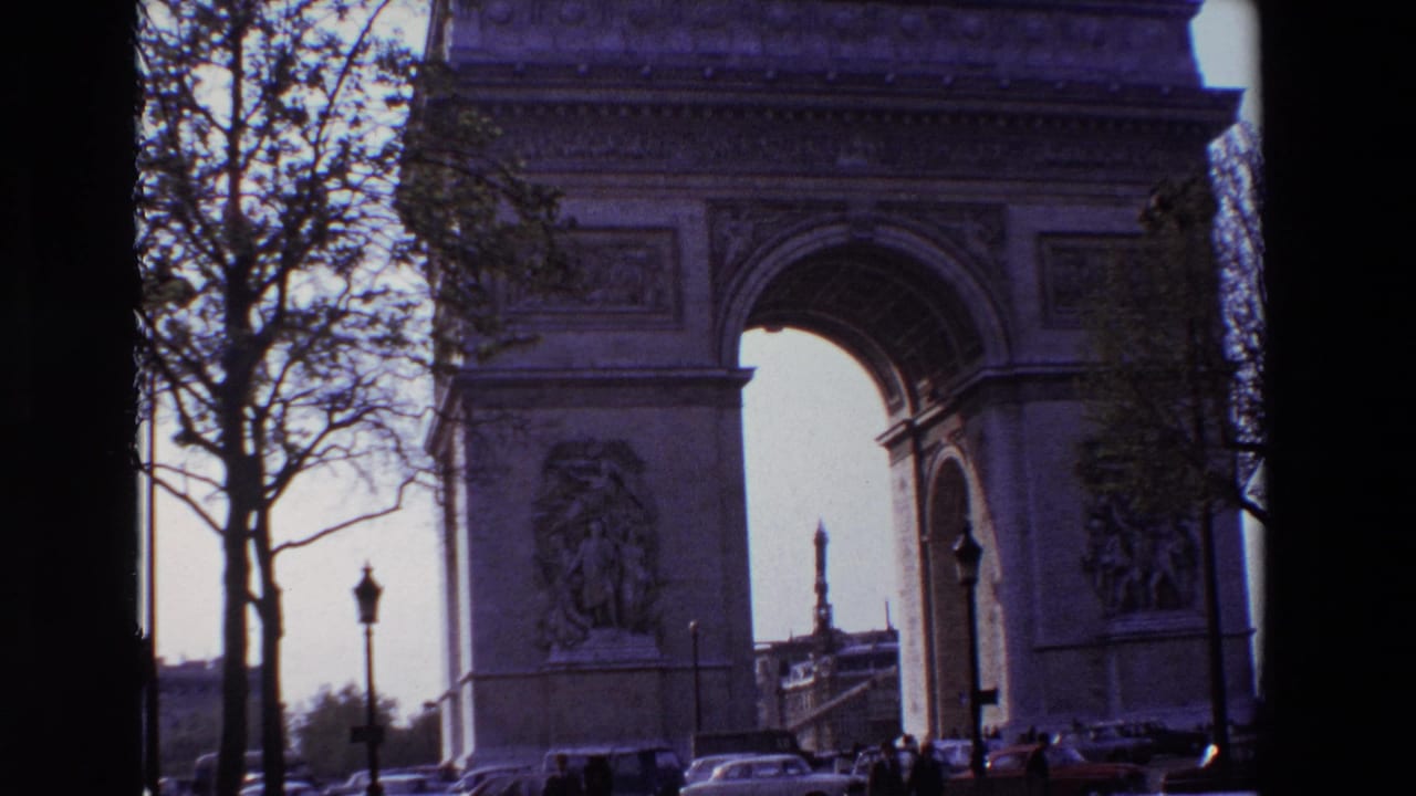 1969 8mm Paris frame showing the Arc de Triomphe from across the street, partially framed by leafless trees with classic cars and small pedestrians below.