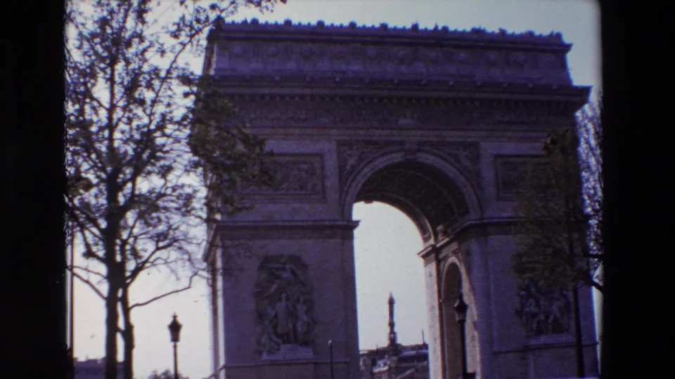 1969 Paris home-movie frame of the Arc de Triomphe with a dark tree trunk at left, a lamppost near the street, and cars visible below the arch.