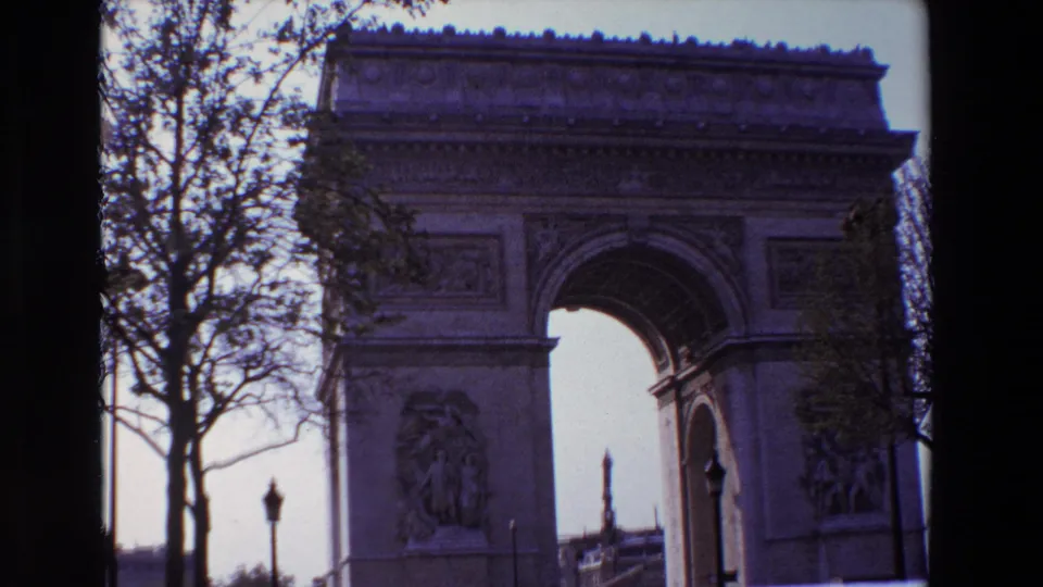 1969 8mm frame from Paris showing the Arc de Triomphe cropped close, with leafless trees, a foreground lamppost, parked or moving cars, and distant pedestrians.