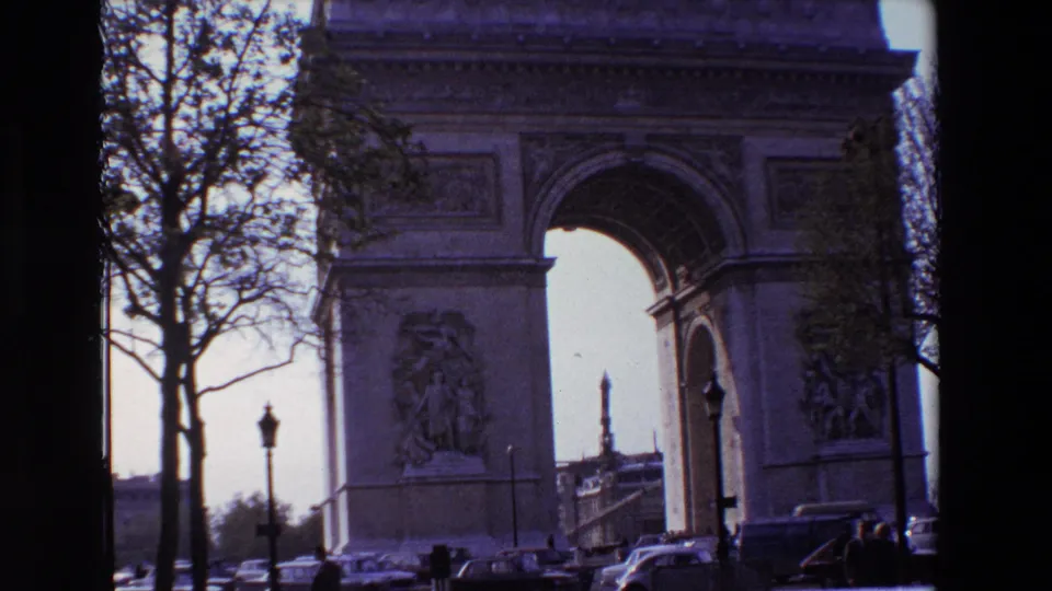 1969 Paris frame showing the Arc de Triomphe through bare branches, with the arch opening, sculptural reliefs, classic cars, and distant pedestrians visible at street level.