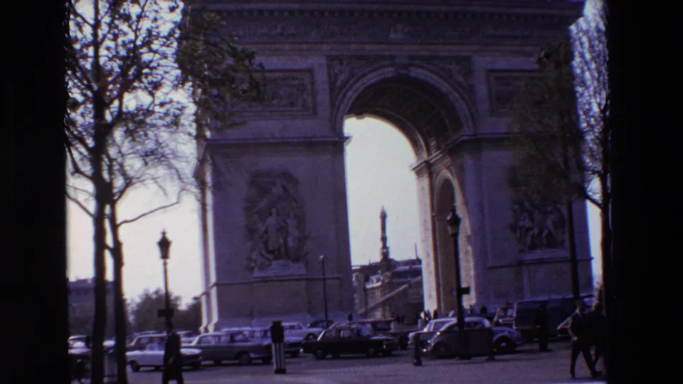 Paris, France in 1969, with the Arc de Triomphe framed by trees and street lamps as pedestrians and cars move through the foreground.