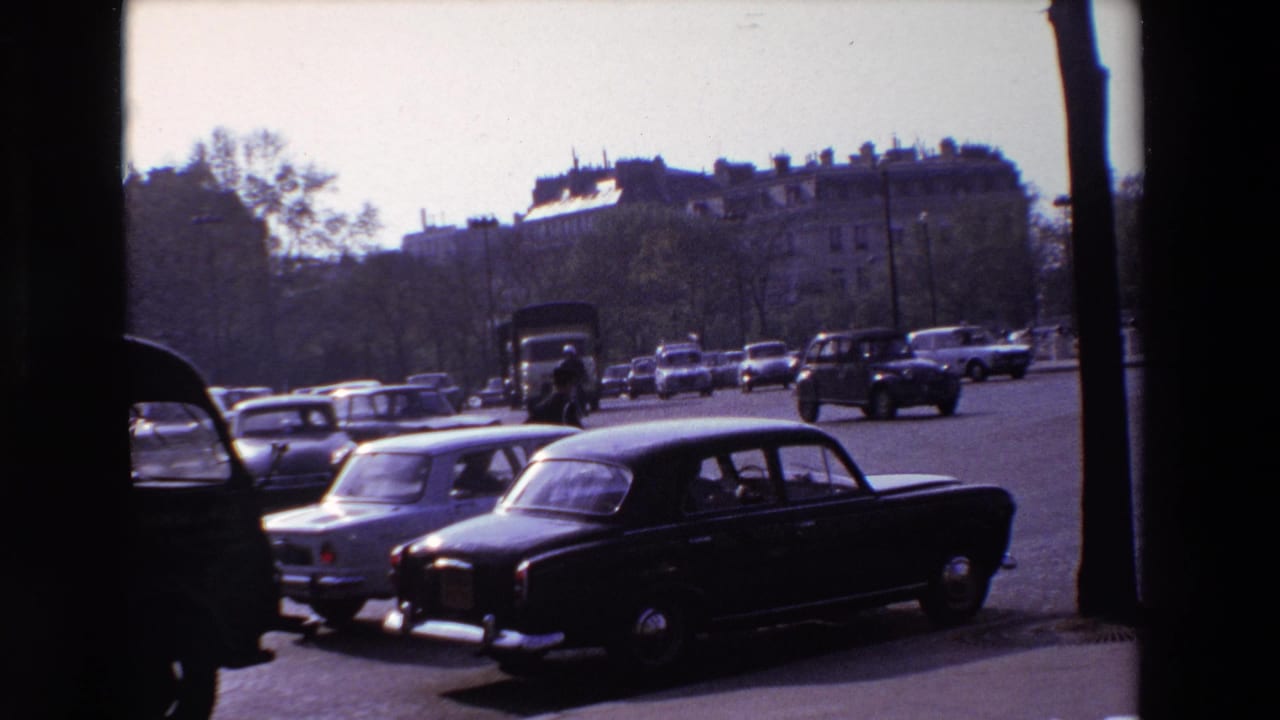 1969 Paris street traffic near the Arc de Triomphe area, with classic cars, larger vehicles, pedestrians, trees, and apartment buildings visible.