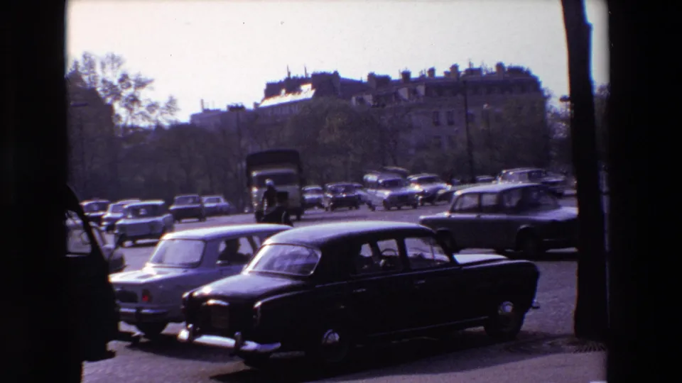 A fixed 8mm view of 1969 Paris traffic shows compact cars and a dark sedan moving past pedestrians, trees, and background buildings.