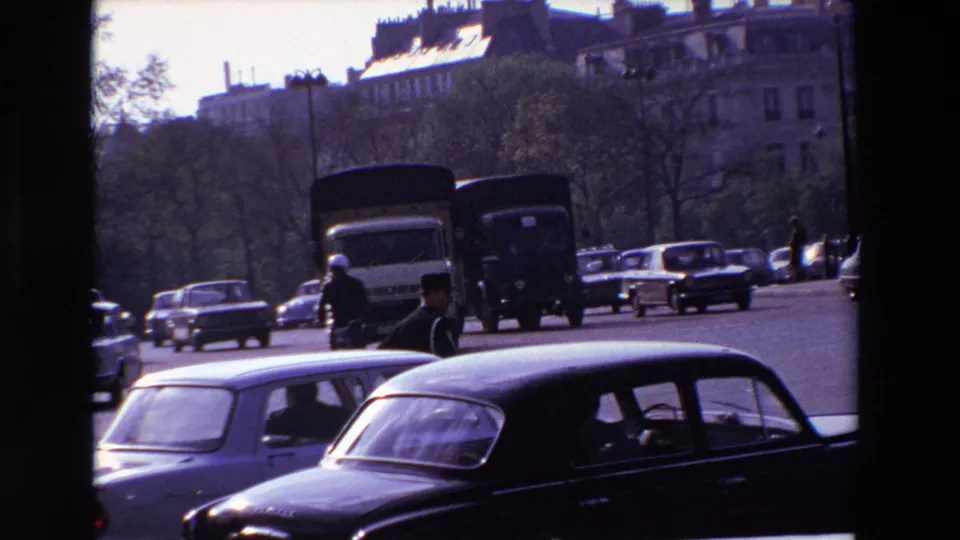 Late-1960s Paris street view with a dark foreground car, larger vehicles, pedestrians, trees, and multi-story buildings beyond the roadway.