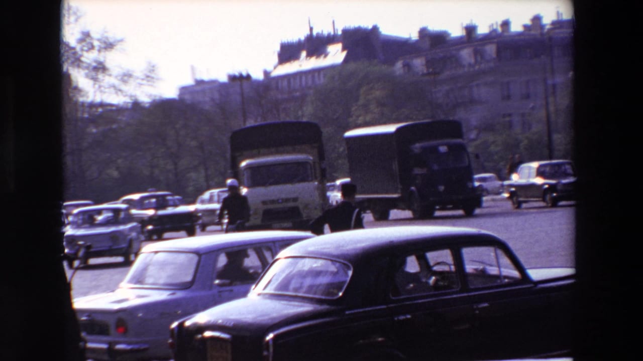 1969 Paris street traffic seen through dark frame edges, with a uniformed policeman directing compact sedans and delivery trucks in bright daylight.