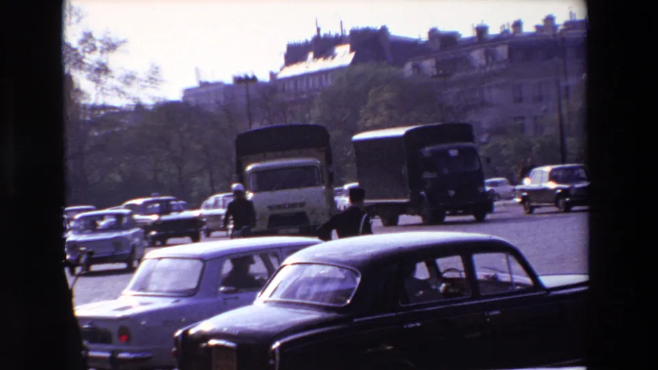 1969 Paris street traffic seen through dark frame edges, with a uniformed policeman directing compact sedans and delivery trucks in bright daylight.
