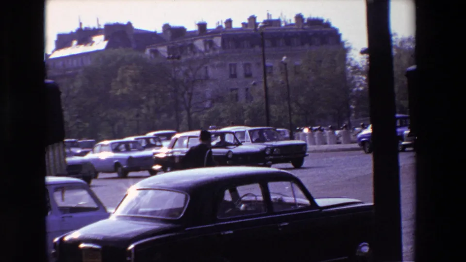 From an obstructed home-movie viewpoint, a uniformed traffic policeman moves among late-1960s Paris street traffic with sedans, trucks, trees, and apartment blocks behind him.
