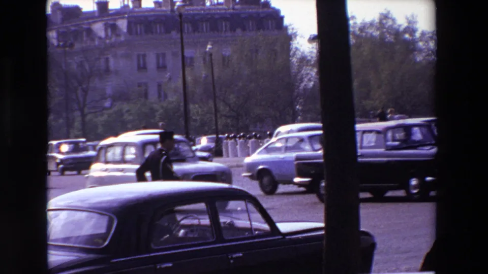 A traffic policeman in Paris stands near a dark sedan as other mid-century cars pass, framed by vertical dark obstructions, trees, and apartment buildings.
