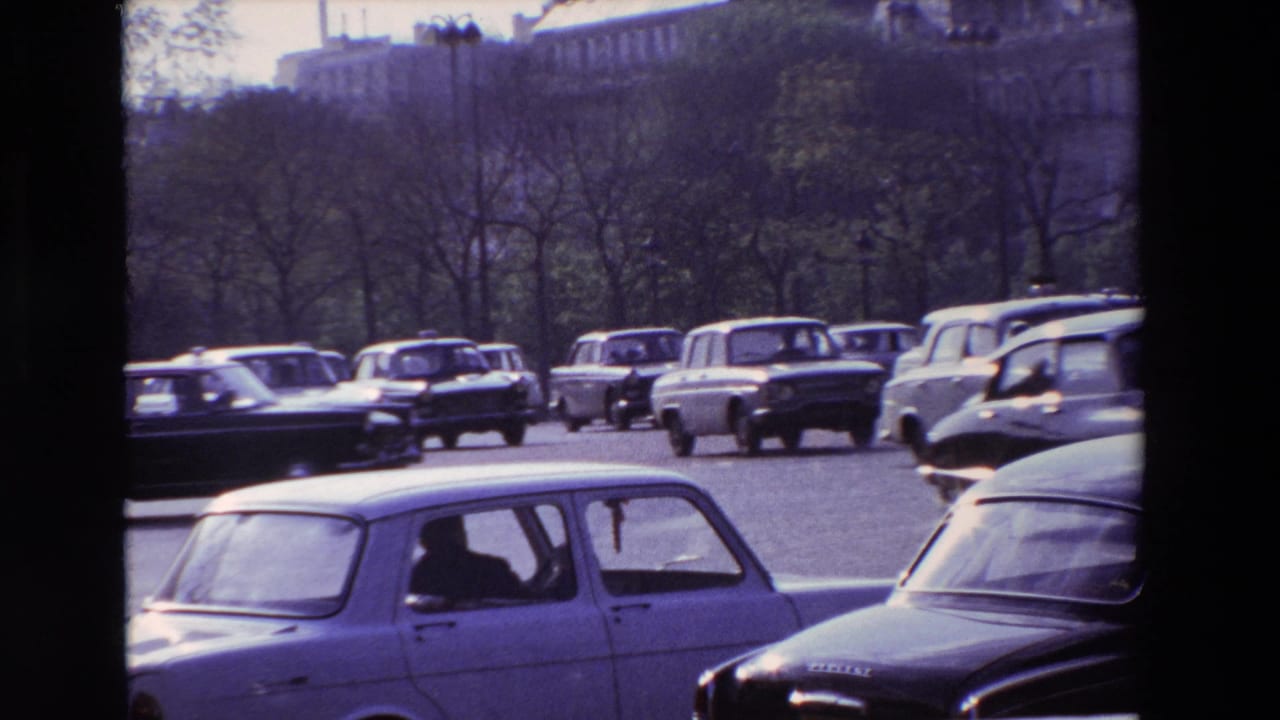 1969 Paris traffic view with compact sedans and station wagons moving across a broad roadway, framed by trees and distant urban buildings.