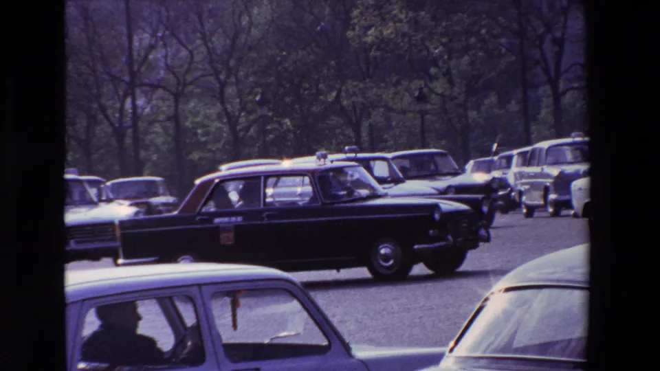 1969 Paris street frame showing a black taxi with a roof sign among classic cars, seen from a handheld 8mm home-movie perspective.
