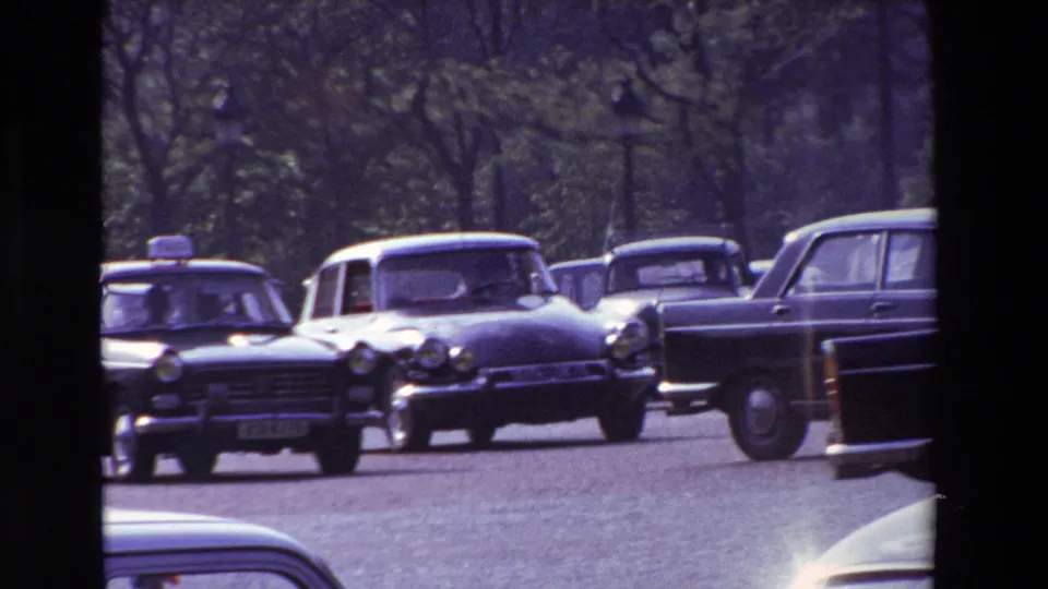 1969 Paris traffic scene with a black taxi, compact cars, and a station wagon moving through a busy roadway under soft, grainy 8mm exposure.
