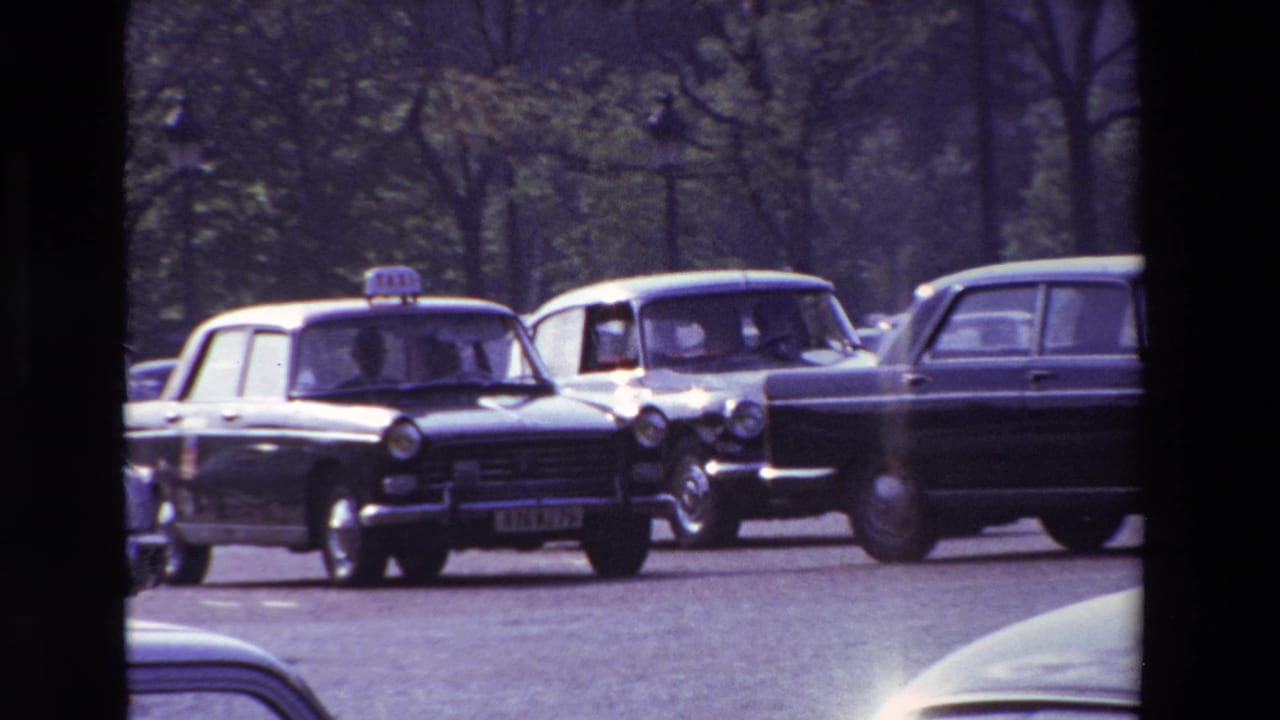 Paris street traffic in 1969 with a black roof-signed taxi, light-colored cars, tree-lined background, and dark vertical foreground obstruction from the filmed viewpoint.