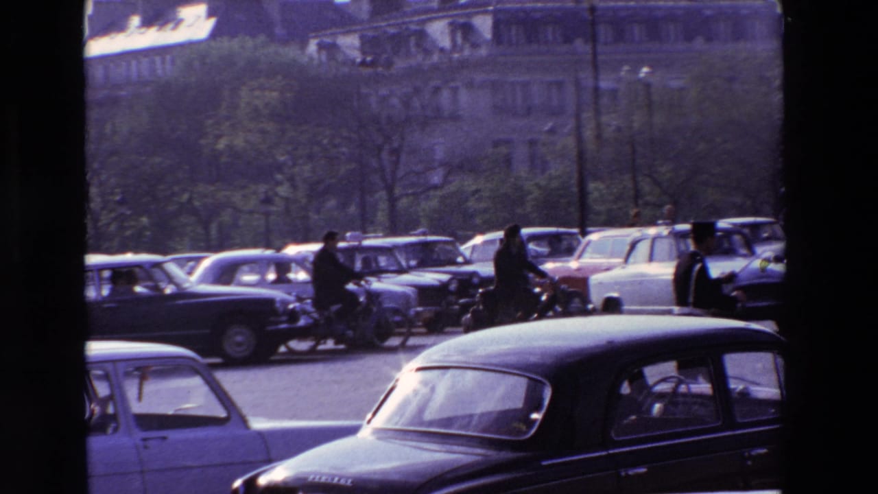 Cars and riders move through traffic on a broad street with buildings and trees behind them.