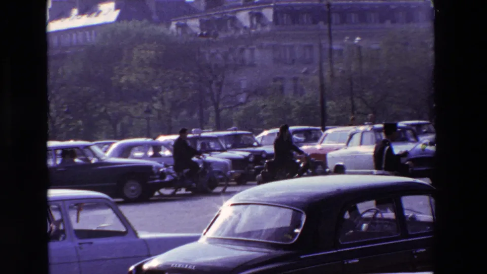 1969 Paris traffic scene with classic automobiles, riders weaving among cars, trees, lampposts, and multi-story buildings in the distance.