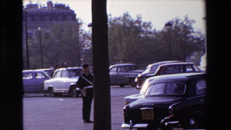 A uniformed traffic or police officer stands beside a Paris roadway in 1969 as classic cars, a black taxi, trees, and stone architecture frame the street scene.