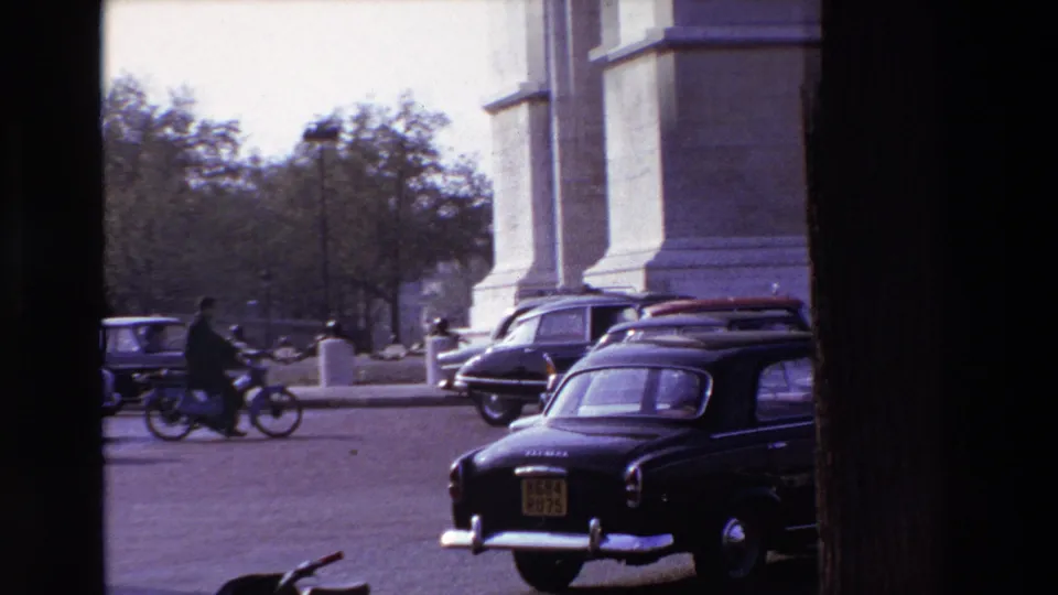 A rider crosses through 1969 Paris street traffic near classic cars, a black taxi, lampposts, trees, and the base of a large stone monument or building.