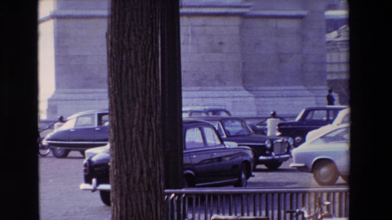 Late-1960s Paris traffic near Place de l'Etoile, with several cars crossing the frame behind foreground tree trunks and the base of a monumental structure behind them.