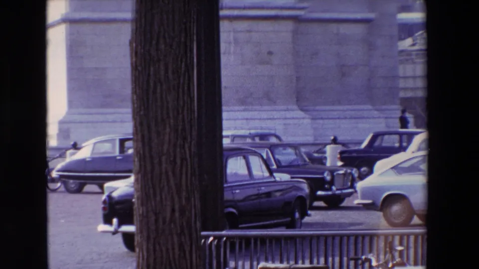Late-1960s Paris traffic near Place de l'Etoile, with several cars crossing the frame behind foreground tree trunks and the base of a monumental structure behind them.