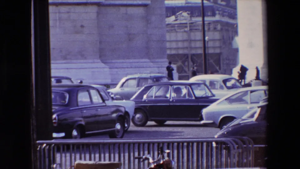 1969 Paris traffic congestion near Place de l'Etoile, showing several mid-century cars, metal barriers, pedestrians, and monumental stonework in soft 8mm color.