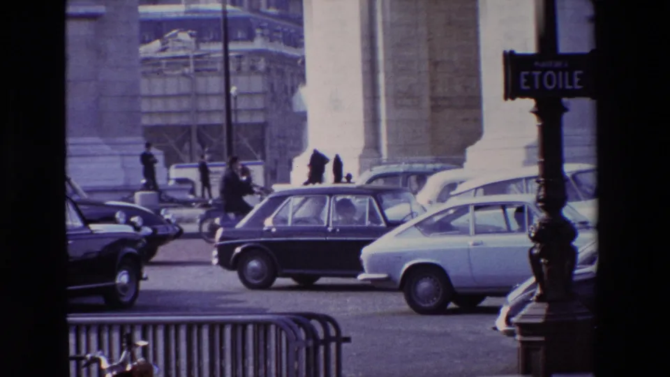Place de l'Etoile street sign visible beside late-1960s Paris traffic, with cars, pedestrians, barriers, tree trunks, and monumental stonework in the background.