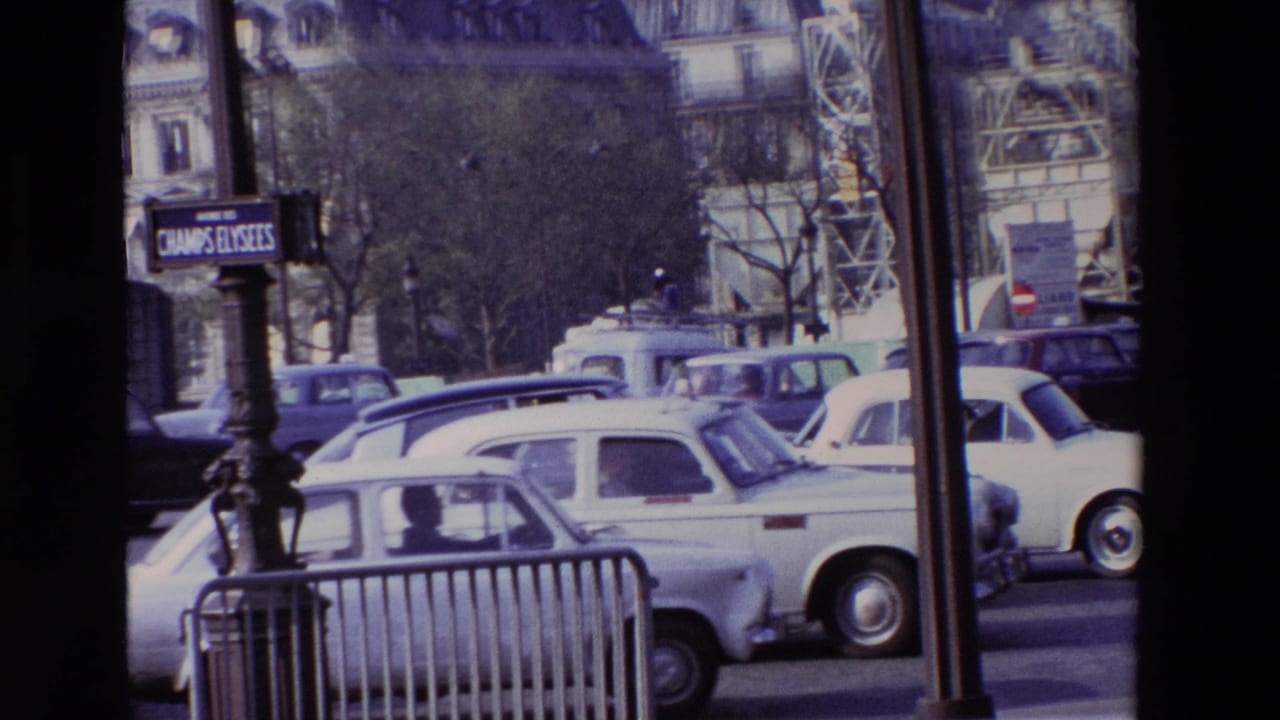 1969 Paris boulevard scene with a Champs-Elysees sign, several older cars, street barriers, trees, and ornate building facades visible behind traffic.