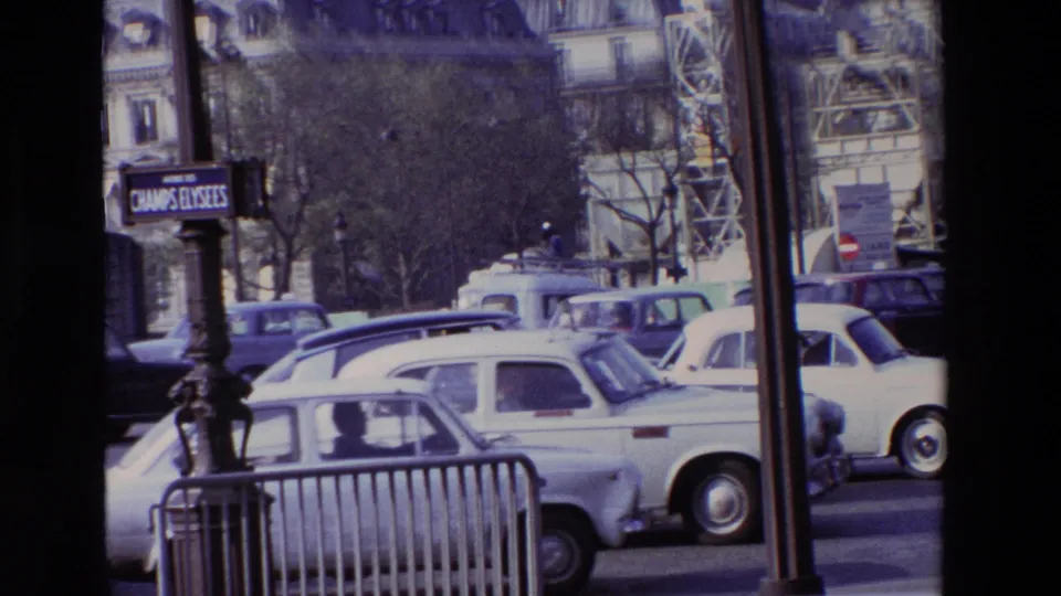 1969 Paris boulevard scene with a Champs-Elysees sign, several older cars, street barriers, trees, and ornate building facades visible behind traffic.