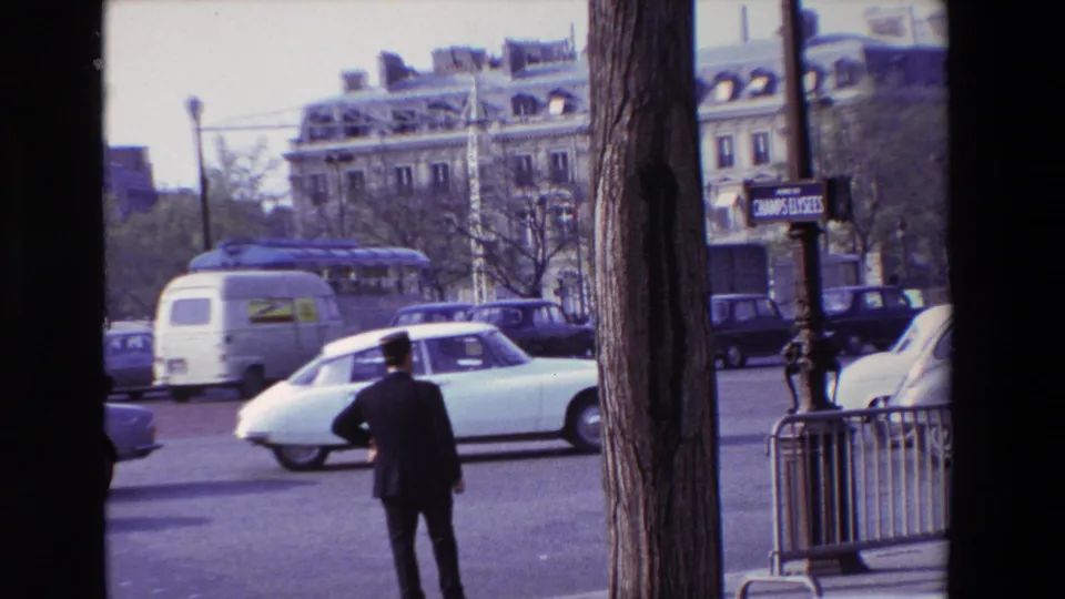 1969 Paris street scene with a man in a dark suit standing near the road while a white van, older cars, trees, barriers, and Champs-Elysees signage appear around the boulevard.