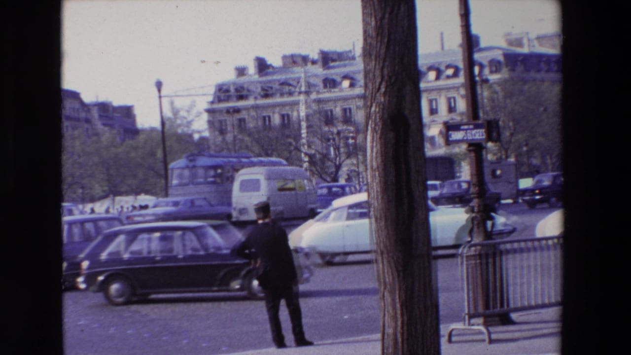 1969 Paris street scene showing a suited man near the curb, classic cars, a bus, street barriers, trees, and ornate buildings beyond the intersection.
