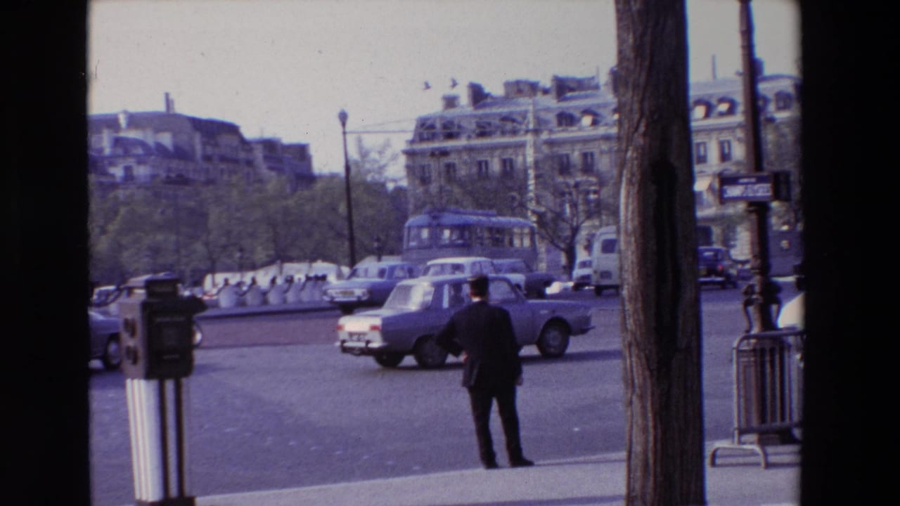 Cars move across a broad intersection while a man stands on the sidewalk near a street fixture.