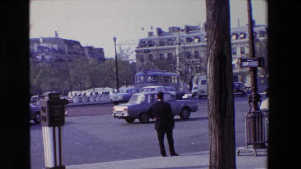 1969 Paris intersection with a suited man facing traffic, several classic cars, a bus in the background, trees, barriers, and upper-floor building facades.