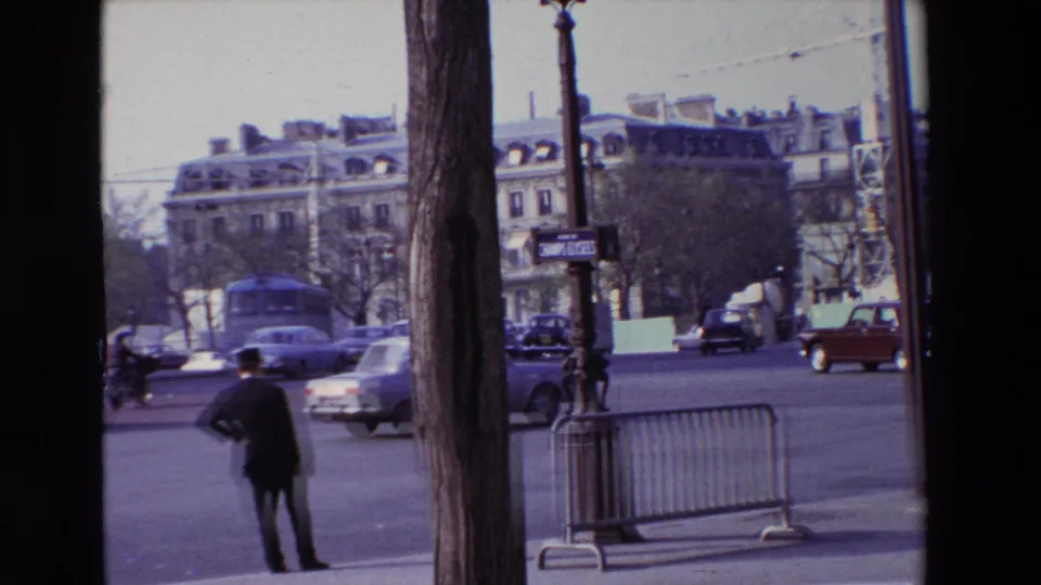 1969 Paris curbside view with a dark-suited man, passing automobiles, a bus, metal pedestrian barriers, street trees, and pale building facades in the background.