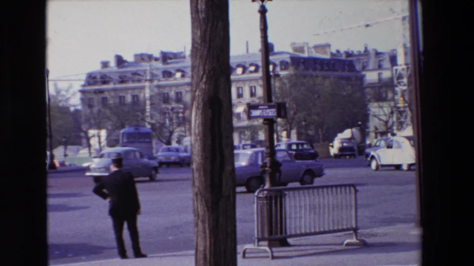 1969 Paris street footage showing a suited man beside barriers, a tree-lined intersection, classic cars, a bus, street posts, and multi-story Parisian buildings.