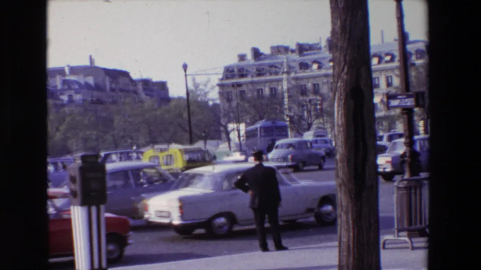 1969 Paris street traffic with classic cars, yellow taxis, sidewalk posts, trees, and a man standing near the curb.