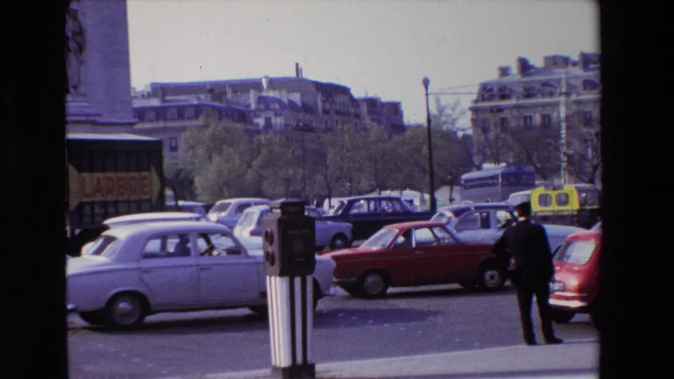 1969 Paris boulevard traffic with a pale sedan, red compact car, yellow taxis, sidewalk fixtures, and a suited pedestrian near the curb.