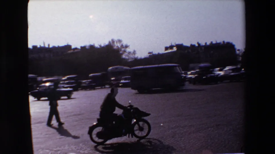 Backlit 1969 Paris traffic scene with a motorcyclist, pedestrians, dark cars, and a bus or van crossing a wide intersection.