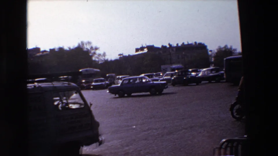 1969 Paris intersection seen in strong backlight, with several cars moving across the boulevard and a foreground vehicle partly cropped by the frame.