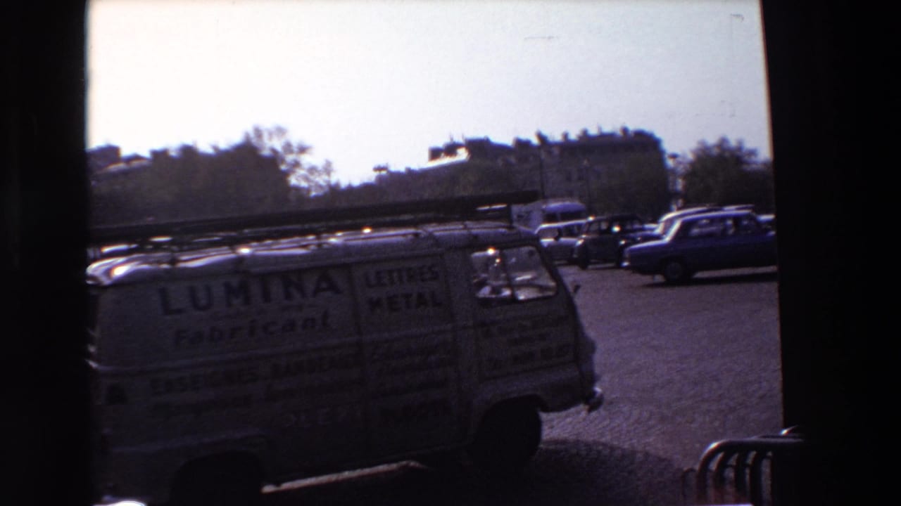 1969 Paris street traffic filmed through a dark frame, showing a commercial van, classic cars, distant buildings, and cobblestone paving.
