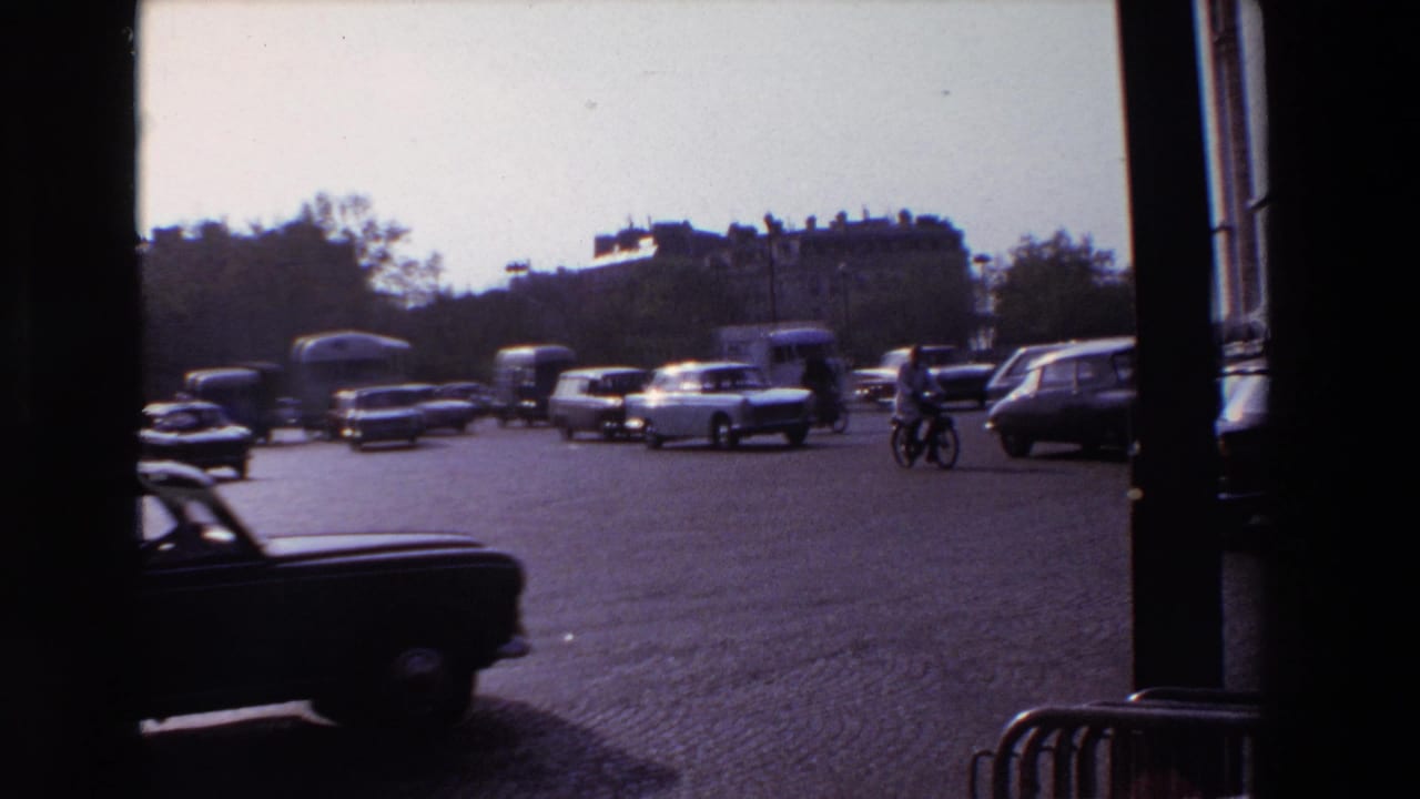 Classic cars and a cyclist move through a broad cobblestone intersection framed by dark vertical posts.