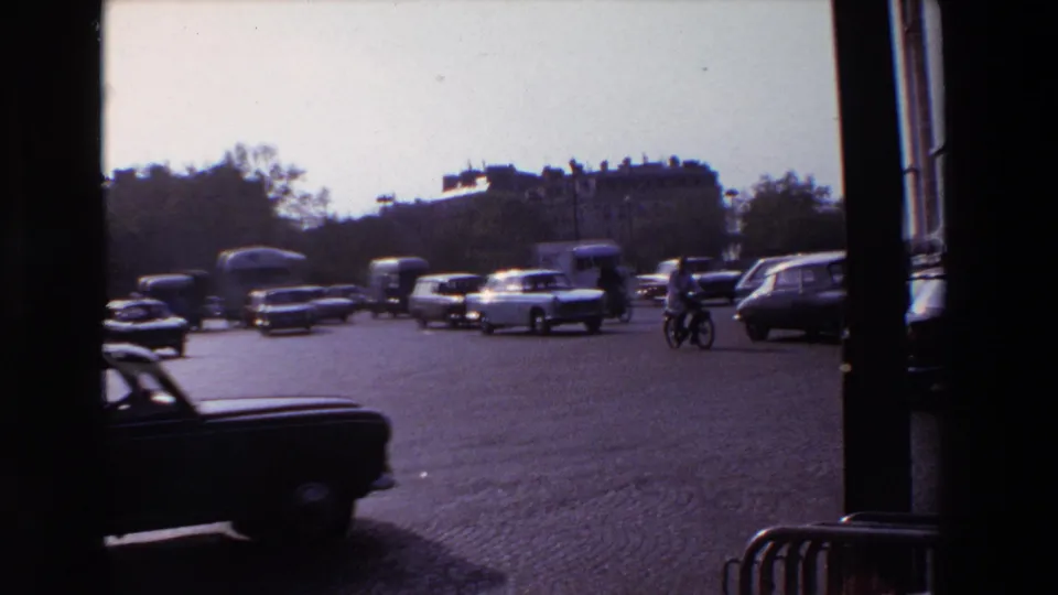 1969 Paris traffic scene with classic cars, a cyclist, cobblestone paving, distant buildings, and dark foreground framing.