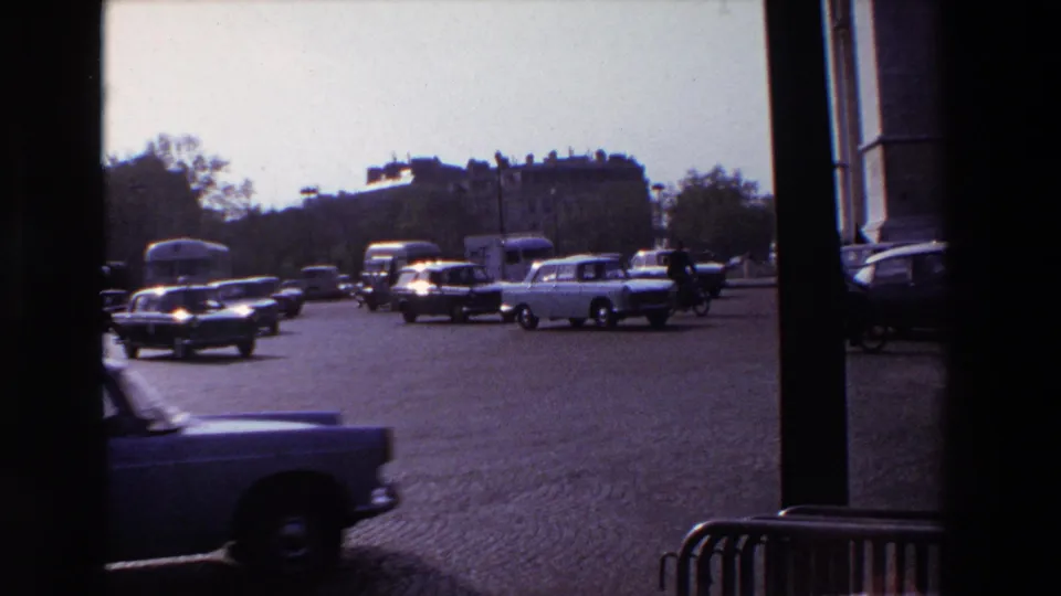 1969 Paris home-movie frame showing cars, a cyclist, cobblestone street surface, dark foreground framing, and hazy city buildings beyond.