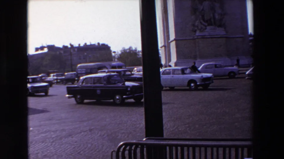 1969 Paris traffic filmed beside a partial stone monument, with classic cars, cobblestone paving, dark foreground posts, and distant buildings visible.