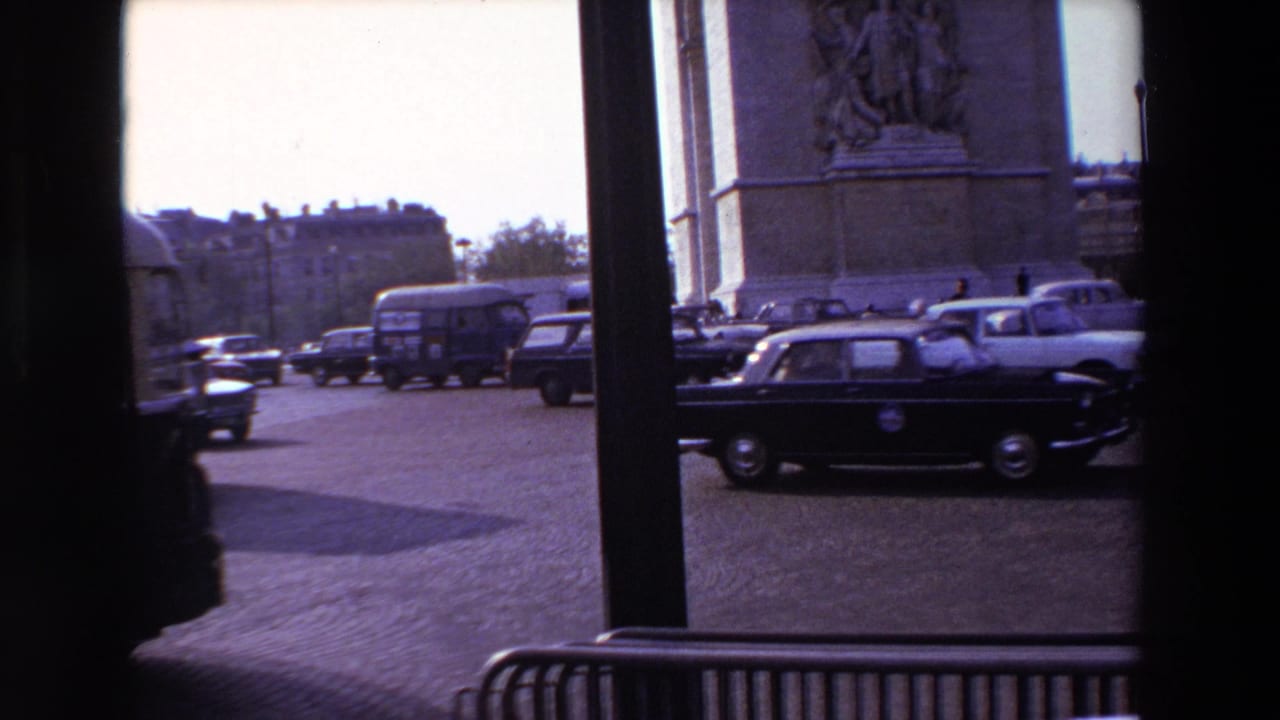 1969 Paris home-movie frame showing cars and a bus moving past the Arc de Triomphe, seen through dark vertical window supports.