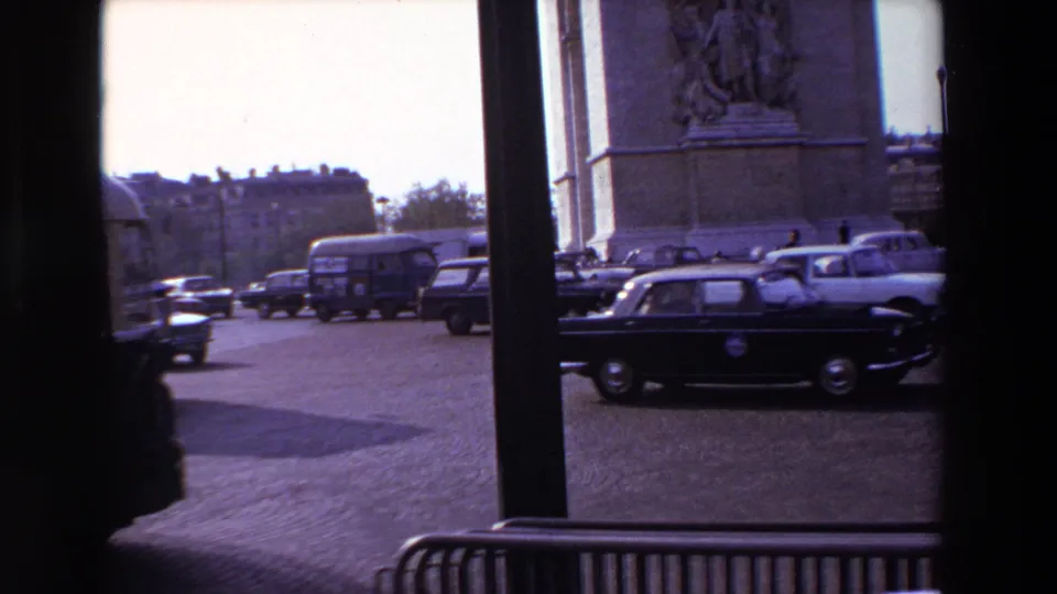 1969 Paris home-movie frame showing cars and a bus moving past the Arc de Triomphe, seen through dark vertical window supports.
