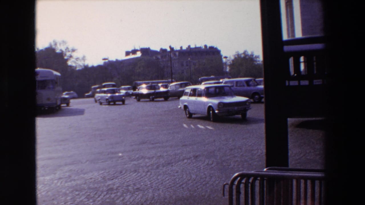 Cars and a white station wagon cross a broad cobblestone roadway near the Arc de Triomphe.