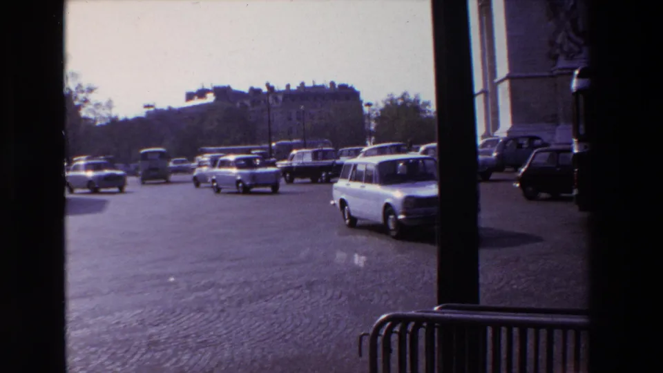 1969 Paris home-movie frame showing a white station wagon, buses, and sedans moving across the Arc de Triomphe traffic area.