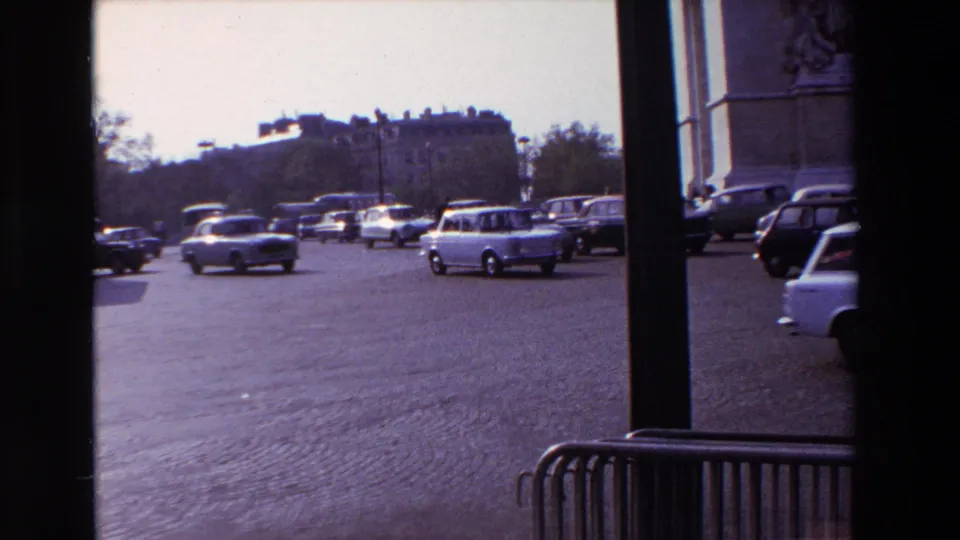 1969 Paris travel frame with classic cars crossing the cobblestone traffic circle near the Arc de Triomphe, partially obscured by window framing.