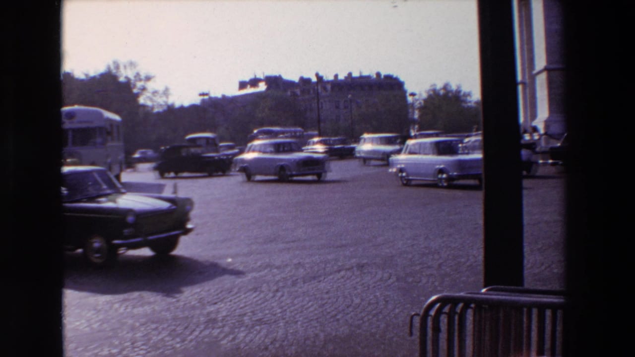 1969 Paris street traffic with mid-century cars and a bus moving across cobblestones, filmed from behind a dark window or doorway frame.