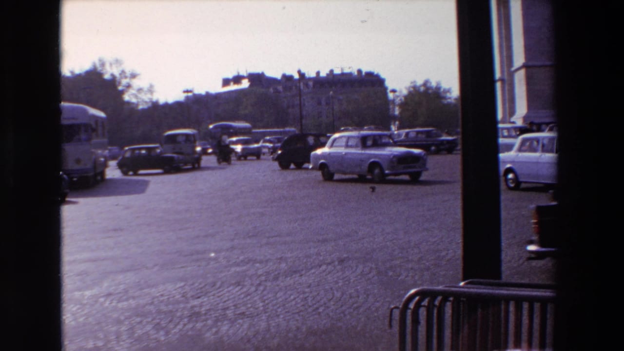 Mid-century vehicles move through a busy Paris traffic scene in 1969, with a bus, sedans, cobblestones, and distant trees visible beyond the window frame.