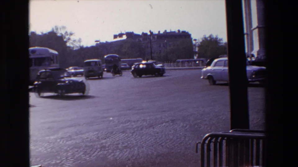 A 1969 Paris traffic view shows sedans and buses crossing a cobblestone roadway, partly obscured by the dark foreground window structure.